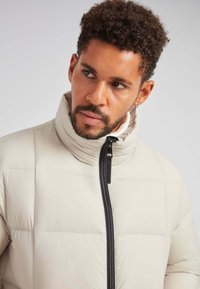 Beige pufferjakke med høj krave, quiltet tekstur og sort lynlås. Glat materiale og tætsiddende form, velegnet til koldt vejr.