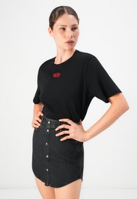 Kvinne iført svart t-skjorte med lite rødt logo og høyt liv svart denimbukse med knapper, hender på hoftene, ser til siden.