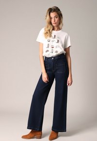 Witte graphic t-shirt met verschillende schoenillustraties; gecombineerd met hoge taille, wijde donkerblauwe jeans en bruine enkellaarsjes.