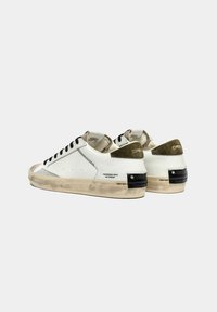 Sneakers bianchi con una texture invecchiata, lacci neri, dettagli in verde oliva e un tallone nero testurizzato. Presentano cuciture visibili e marchi.