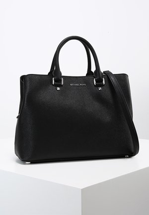 Sac à main en cuir texturé noir Michael Kors avec double poignée et bandoulière amovible, reposant sur un piédestal blanc.