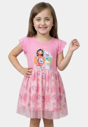 Jeune fille souriante portant une robe rose avec des personnages de dessin animé sur le corsage et des empreintes de pattes sur la jupe, posant avec une main sur la hanche.