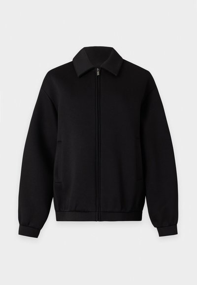 Veste noire zippée avec manches longues, col, poches latérales et poignets et ourlet côtelés, présentée sur un fond clair uni.