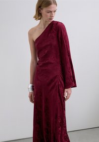 Robe de soirée bordeaux asymétrique en tissu doux avec un motif subtil, présentant une longue manche drapée et un détail froncé sur le côté.