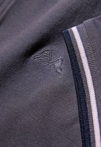 Grauer Stoff mit einem dezenten, in das Gewebe eingestickten Logo. Auf einer Seite mit einem dunkelblau-weiß gestreiften Rand akzentuiert.