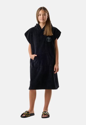 Girl wearing a black hooded towel dress with a front pocket and black slides featuring a yellow duck pattern, standing against a plain background.