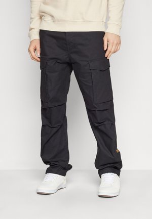Homme portant un pantalon cargo noir avec des poches à rabats et des baskets blanches, debout devant un fond blanc uni.