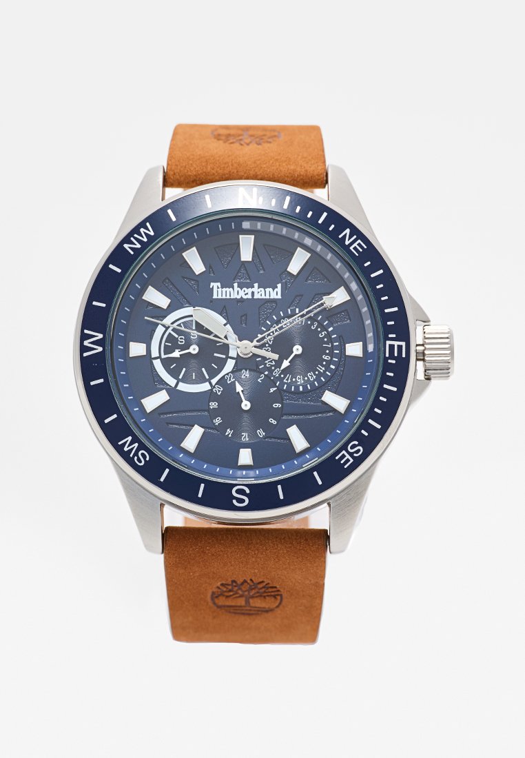 Reloj de metal plateado con un dial azul marino, correa de cuero marrón, marcadores de hora plateados y tres sub-diales. Logotipo de Timberland grabado en la correa.