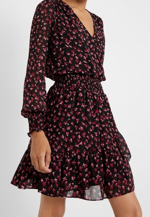 Woman wearing black long-sleeve dress with pink floral pattern, elastic smocked waist, and ruffled hem, standing against white background.