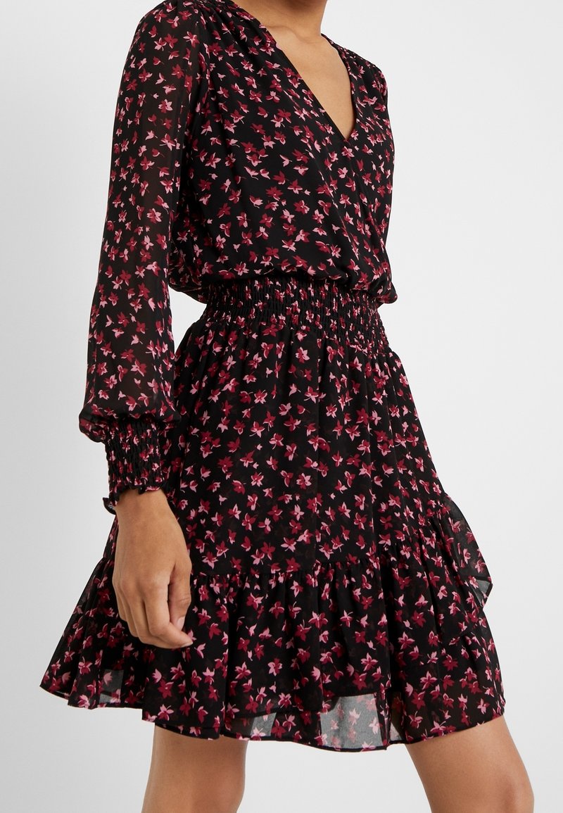 Žena v černých šatech s dlouhým rukávem s růžovým květinovým vzorem, elastickým nařaseným pasem a volánkovým lemem, stojící na bílém pozadí.