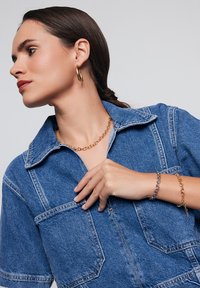 Kurtka dżinsowa z kołnierzem w kolorze niebieskim i przednimi kieszeniami. Na szyi złote i srebrne naszyjniki z łańcuszków oraz kolczyki koła.