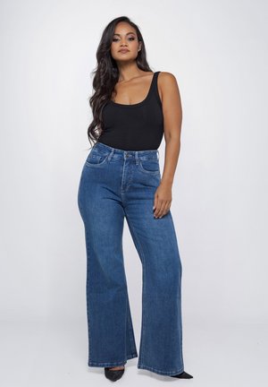 Vrouw staat zelfverzekerd en draagt een zwarte mouwloze top, hoge wijd uitlopende blauwe jeans en zwarte puntige schoenen tegen een effen achtergrond.