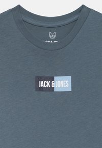 Dunkeltürkises T-Shirt aus weichem Stoff mit einem rechteckigen Logo in Schwarz und Hellblau sowie dem Text "JACK & JONES" in Weiß.