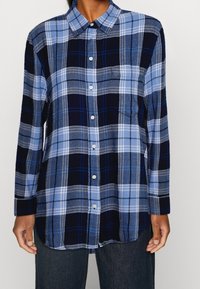 Chemise à manches longues à carreaux dans des tons de bleu, marine et gris, avec un devant à boutons, un col et une poche sur le côté gauche.