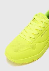 Sneaker lumineux jaune fluo avec une tige en synthétique lisse, des détails perforés, des lacets plats et une semelle intérieure rembourrée. Nom de la marque visible sur la languette.