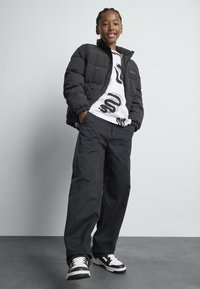 Zwarte pufferjack met rits, wit graphic t-shirt, zwarte broek en zwart-witte sneakers. De jas heeft een hoge kraag en geribbelde manchetten.