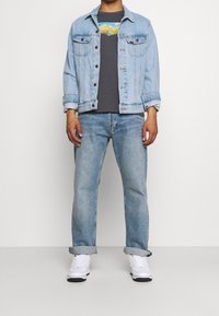 Persona con chaqueta de mezclilla azul clara, camiseta gráfica negra, jeans azul claros con puños y zapatillas blancas, de pie contra un fondo plano.