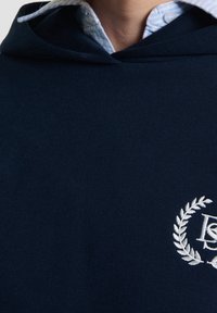 Sudadera con capucha azul marino con un acabado texturizado, que presenta un logo bordado en blanco y un patrón de laurel en el pecho. Cuello azul claro con botones visible.