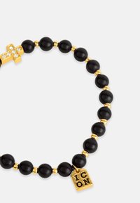 Juodas karoliukų apyrankė su auksinėmis detalėmis, kryžiaus akcentu, puoštu cirkoniukais, ir žyma su įrašu „ICON“. Bendras paviršius lygus.