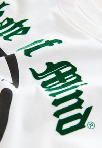 T-shirt blanc avec un texte vert en relief dans une police ludique. Le tissu semble doux, avec une texture lisse et sans motifs apparents.
