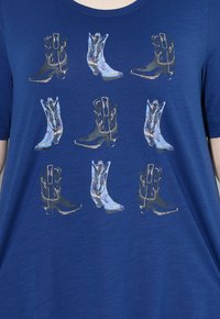 Modrá tričko s opakujícím se grafickým vzorem kovbojských bot v zlaté a stříbrné barvě, uspořádaným do mřížkového vzoru na látce.