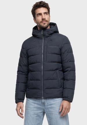 Mann trägt eine dunkelblaue gesteppte Jacke mit Kapuze und Reißverschluss, kombiniert mit hellblauen Jeans, vor einem schlichten Hintergrund stehend.