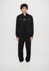 Svart zip-jacka med ståkrage, prydd med gyllene logodetaljer, i kombination med matchande svarta byxor och vita sneakers.