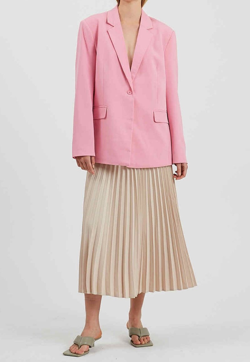 Femme portant un blazer rose sur une jupe midi plissée beige et des sandales à talons vertes à bout ouvert, debout devant un fond clair uni.