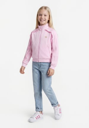 Chica sonriente con chaqueta rosa con cremallera, vaqueros azul claro remangados y zapatillas blancas con rayas rosas, de pie sobre un fondo blanco.
