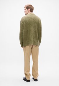 Groen, gebreide cardigan met textuur en een ontspannen pasvorm, gecombineerd met lichtbeige broek en zwarte schoenen, gezien vanaf de achterkant.