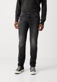 Jean en denim foncé avec une coupe slim, présentant une légère usure, cinq poches, un bouton en cuivre, associé à des baskets en toile noires.
