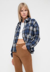 Blå og svart rutete flanellskjorte med krage, knappelukking og sidelomme, kombinert med brune kordfløydbukser.