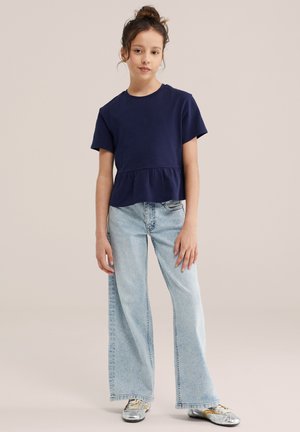 Mladá osoba stojaca s uvoľnenými rukami, oblečená v námornícky modrom tričku s krátkym rukávom, svetlomodrých širokých rifliach a strieborných teniskách.