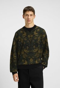 Giovane uomo che indossa un maglione a fantasia floreale verde scuro e marrone con uccelli, abbinato a pantaloni neri, in piedi contro uno sfondo neutro.