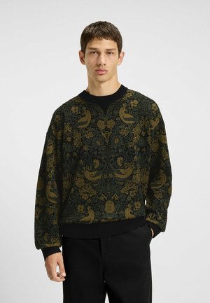 Junger Mann in einem dunkelgrünen und braunen Sweatshirt mit floralem Muster und Vögeln, kombiniert mit schwarzen Hosen, vor einem einfarbigen Hintergrund stehend.