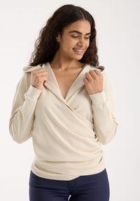 Beige huppari, jossa on ristikkäinen muotoilu, valmistettu sileästä kankaasta, jossa on pitkät hihat ja rutistettuja yksityiskohtia vyötäröllä.