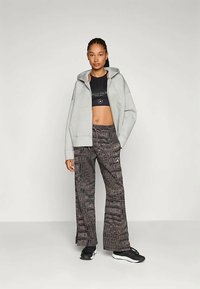 Šedá mikina na zip přes černou crop top, kombinováno s tmavě vzorovanými širokými kalhotami a černými sportovními botami.