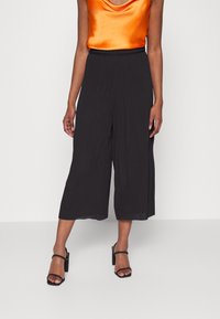 Frau, die lockere schwarze Cropped-Hosen, ein oranges satinähnliches ärmelloses Oberteil und schwarze Sandalen mit Absatz und Knöchelriemen trägt, steht vor einem weißen Hintergrund.
