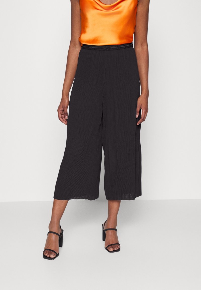 Frau, die lockere schwarze Cropped-Hosen, ein oranges satinähnliches ärmelloses Oberteil und schwarze Sandalen mit Absatz und Knöchelriemen trägt, steht vor einem weißen Hintergrund.