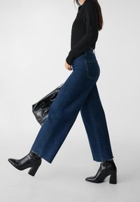 Woman in dark blue wide-leg jeans, black long-sleeve top, black ankle boots, carrying black studded handbag, walking against plain background.