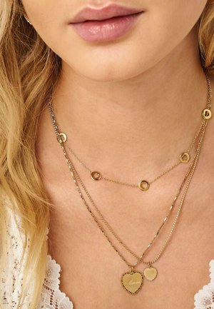 Gros plan sur le cou d'une femme avec des colliers en or superposés, comprenant des pendentifs en forme de cœur et des breloques circulaires, sur un tissu en dentelle.