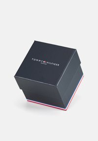 Schwarze Uhrenbox aus Kunststoff mit glänzender Oberfläche. Verfügt über das weiße Logo "TOMMY HILFIGER" und einen rot-weiß-blauen Streifenakzent an der Basis.