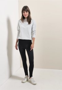 Felpa grigia con polsini e scollatura a costine abbinata a leggings neri. Il modello indossa sneaker bianche davanti a uno sfondo neutro.