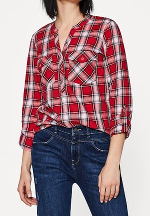 Kobieta ubrana w czerwoną i białą koszulę w kratę z długim rękawem i dwoma kieszeniami na piersi oraz ciemnoniebieskie dżinsy, dłonie rozluźnione wzdłuż ciała.