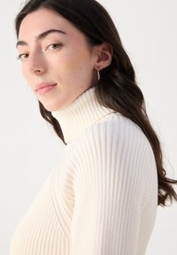 Krémový, skladaný rolák s vysokým golierom a priliehavým strihom. Modelka nosí strieborné kruhové náušnice. Hladká textura a upravený fit.