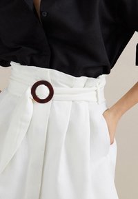 Parte centrale di una donna che indossa una camicia nera con bottoni infilata in pantaloni bianchi a vita alta con una cintura larga e una fibbia scura rotonda.