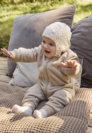 Beige fleece-dragt med lynlås og ribbede manchetter; mønstret strikket hue; sidder på teksturerede puder, smilende med armene løftet.