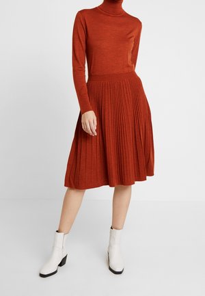 Kobieta ubrana w długą sukienkę z golfem i plisowanym dołem w kolorze rdzawym, w połączeniu z białymi botkami, stojąca na jednolitym tle.