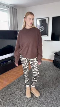 Mujer de pie en una sala de estar vistiendo un suéter marrón, leggings con rayas de cebra y botines beige.