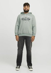 Sudadera verde claro con texto "Jack & Jones Surfer's Club" en color marino. Combinada con jeans de denim oscuro y zapatillas negras con suelas blancas.